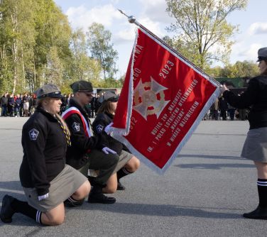 po prawej kobieta pochyla sztandar do przodu, po lewej trzy osoby dwie kobiety i jeden mężczyzna klęczą przed sztandarem, w...