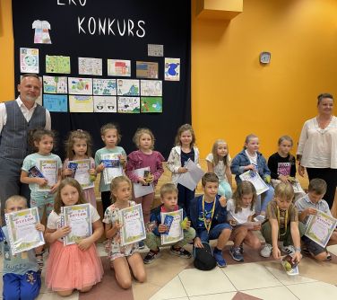grupa dzieci i dwoje dorosłych pozują do zdjęcia w auli szkolnej w tle na ścianie czarna płachta na niej wiszą prace i...