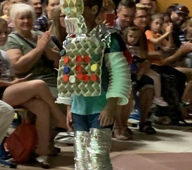 aula szkolna, pokaz mody, chłopiec przebrany za robota z wytłaczanek po jajkach, na rękach plastikowe rury, na nogach...