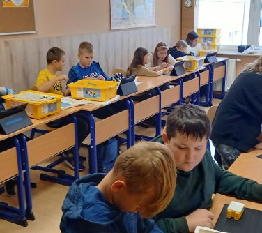 sala lekcyjna, przy stolikach połączonych jeden obok drugiego siedzą dzieci , na stołach pudełka z klockami, obok tablety