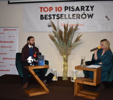 Kobieta i mężczyzna siedzą przy stoliku na scenie, kobieta mówi do mikrofonu