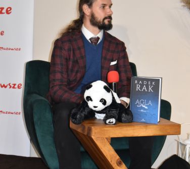 mężczyzna z długimi włosami i brodą siedzi na zielonym fotelu z mikrofonem w ręce, przed nim stolik z książką i pluszową...