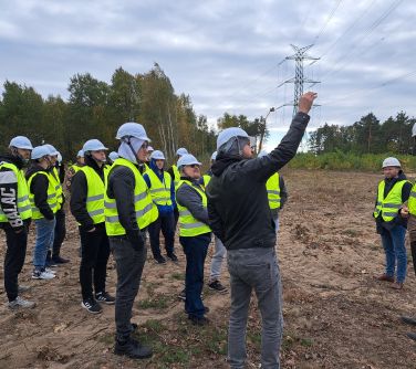 grupa osób stoi na piachu w kaskach i kamizelkach odblaskowych, skierowani na prawo, mężyczyzna z boku wskazuje coś dłonią...