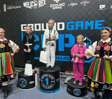 trzy dziewczynki stoją na podium, po obu stronach kobiety ubrane w stroje ludowe, w tle baner