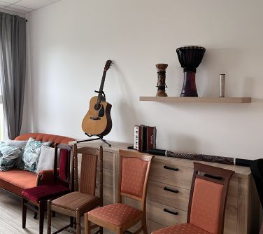 pokój, łóżko, lrzesła za krzesłami komoda na nich instrumenty, nad komoda wisząca półka na niej instrumenty