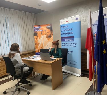 pokój, biurko za biurkiem na krześle siedzi kobieta, przed nią na krześle do biurka siedzi druga kobieta, w tle trzy...