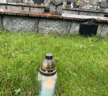 zapalony znicz z szarfą niebiesko-żółtą stoi na trawie, w tle pomniki