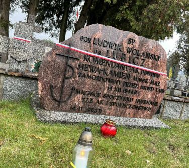 zapalony znicz z szarfą niebiesko-żółtą stoi na trawie za nim czerwony znicz, w tle kamień ś.p. por Ludwik Wolański ps...