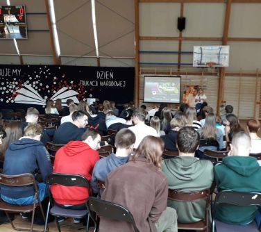 grupa nastolatków siedzi na krzesłach na hali sportowej, na przodzie baner ślubujemy Dzień Edukacji narodowej, rkran, rolap...