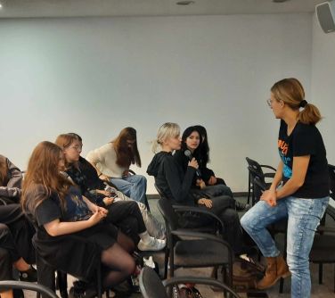 aula, krzesła, na nich osoby, na przodzie kobieta usiadła na tyle krzesła, do mikrofonu przemawia nastolatka siedząca na...