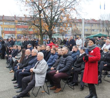 zaproszeni goście siedzą na krzesełkach, w tle stoja mieszkańcy i namioty, po środku drzewo