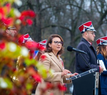kobieta przemawia stoją przy mównicy, za nią kilka osób z orkiestry, na przodzie po lewej kwiaty