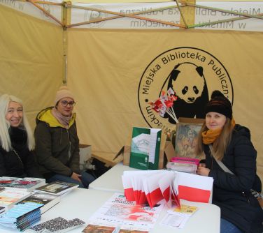 trzy kobiety siedza pod namiotem miejska biblioteka publiczna, obok nich stoły z publikacjami