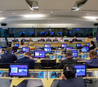 osoby siedzą w słuchawkach przy podłuznych stołach, na stoąłch laptopy, na przodzie przy podłużnym stole prowadzący