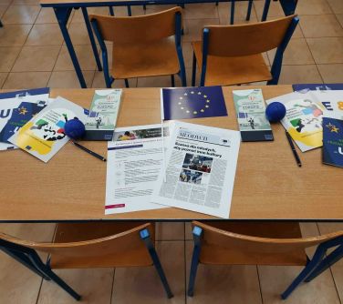 stół i krzesła w sali lekcyjnej, na stole broszury, flaga, gadżęty o tematyce Unii Europejskiej