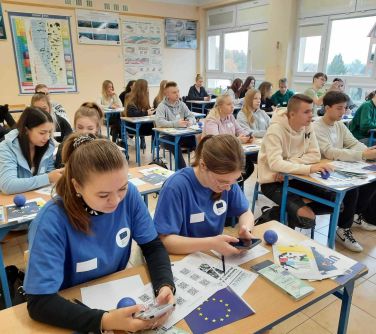 młodzież siedzi w łakwach szkolnych, dwie nastolatki po lewej stronie w niebieskich koszulkach sprawdzają coś w telefonach