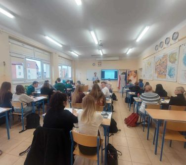 zdjęcie sali lekcyjnej, w ławkach szkolnych siedzi młodzież, na przodzie mężczyzna i ekran z wyświetlana treścią