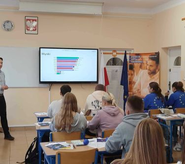 młodzież szkolna siedzi w ławkach w sali lekcyjnej, na przodzie ekran z wyświetlaną treścią, po prawej stronie stoi roll-up