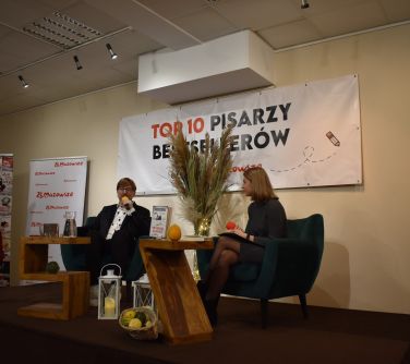 na podwyższeniu fotele i dwa stoliki, na fotelach siedzi mężczyzna i kobieta. mężczyzna przemawia. pomiędzy nimi wysoki...