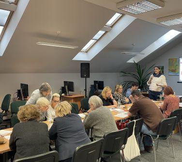 osoby siedzą naokoło złączonych stołów, kobieta na przodzie przemawia