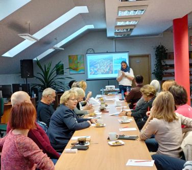 osoby siedzą naokoło złączonych stołów, kobieta na przodzie przemawia, za kobietą ekran z wyświetlaną treścią