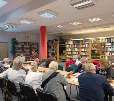 osoby siedzą naokoło złączonych stołów, w tle regały z książkami