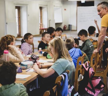 Sala z lustrami. Duża grupa dzieci siedzi na krzesłach naokoło żłączonych stołów, nad nimi pochyla się mężczyzna