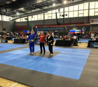 Hala sportowa. Na piankowej macie obok siebie stoi trzech mężczyzn. Mężczyzna w srodku podnosi do góry rękę stojącego po...