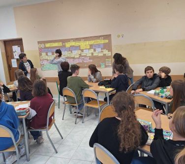 Sala. Dzieci siedzą przy kwadratowych stołach, na przodzie tablica z zaczepionymi, kolorowymi kartkami
