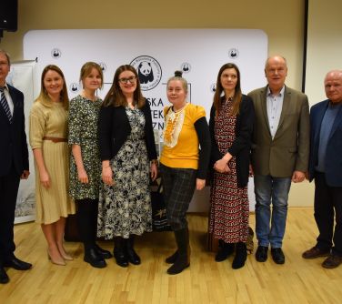 Aula w bibliotece. Osiem osób pozuje do zdjęcia. W tle baner Miejskiej Biblioteki Publicznej
