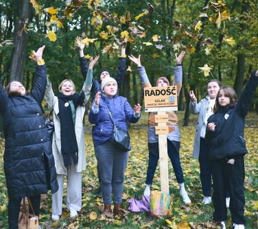 Park. Siedem osób podrzuca liście do gówy. Po środku drewniana tabliczka z napisem radość