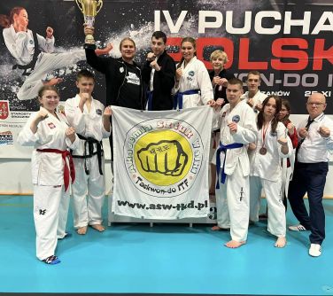 Hala sportowa. Jedenaście osób pozuje do zdjęcia trzymając na przodzie flagę Akademii Sztuk Walki. Cztery osoby po środku...