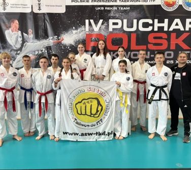 Hala sportowa. Grupa osób pozuje do zdjęcia trzymając na przodzie flagę Akademii Sztuk Walki