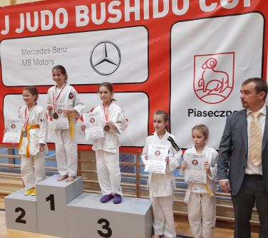 Hala sportowa. Na podium trzy dziewczynki, obok dwie dziewczynki i mężczyzna. Dziewczynki trzymają dyplomy