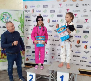 Hala. Dwie dziewczynki na podium trzymają w dłoniach dyplomy. Po lewej stoi mężczyzna