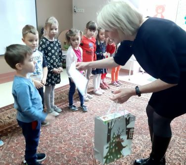 Sala przedszkolna. Siedmioro dzieci, przed nimi kobieta schylająca się do pierwszego chłopca. Trzyma torbę prezentową jedną...