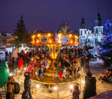 Dwór. Zima. Osoby przechadzają się po placu. Na środku rozświetlona karuzela