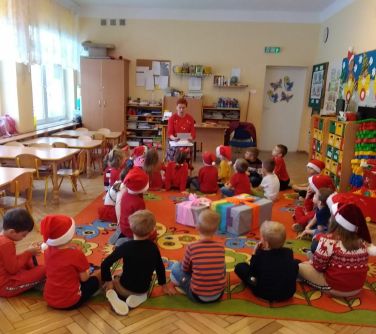 Sala przedszkolna. Grupa dzieci siedzi w czapkach mikołajowych na dywanie. Po środku dwa duże prezenty. Na przodzie siedzi...