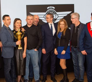 Pomieszczenie. Dziewięć osób pozuje od zdjęcia. Mężczyzna po lewej trzyma puchar