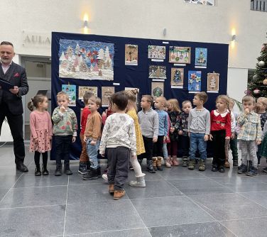 Aula. Mężczyzna i grupa dzieci stoi przed planszą z rozwieszonymi pracami plastycznymi