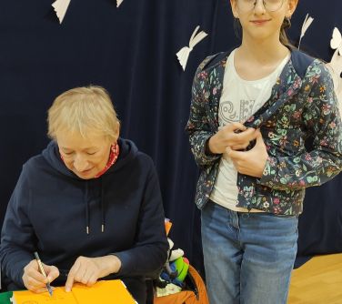 Sala gimnastyczna. Kobieta siedzi i podpisuej przy stoliku książkę. Obok stoi dziewczynka