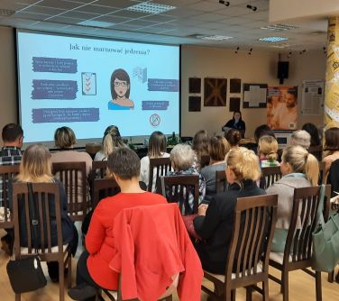 Aula. Na krzesłąch siedzą osoby. Na przodzie kobieta opowiada. Na ekranie wyświetlane informacje.