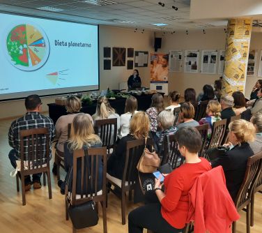 Aula. Osoby siedza na krzesłach. Na przodzie kobieta. Na ekranie wyświetlana treść
