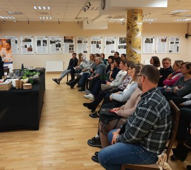 Aula. Grupa osób siedzi na krzesłach. Na przodzie przemawia kobieta stojąc przed stołem