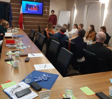 Sala konferencyjna. Na krzesłach przy stole ułożonym w podkowe siedzą osoby. Na przodzie przemawia mężczyzna
