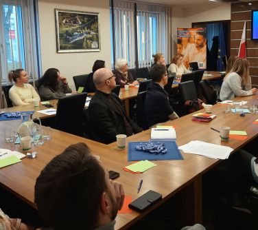 Sala konferencyjna. Na krzesłach przy stole ułożonym w podkowe siedzą osoby.