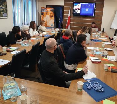 Sala konferencyjna. Na krzesłach przy stole ułożonym w podkowe siedzą osoby.