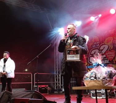 Na oświetlonej scenie kobieta z lewej, na przodzie mężczyzna przemaiwa do mikrofonu. Obok niego stolik z upominkami
