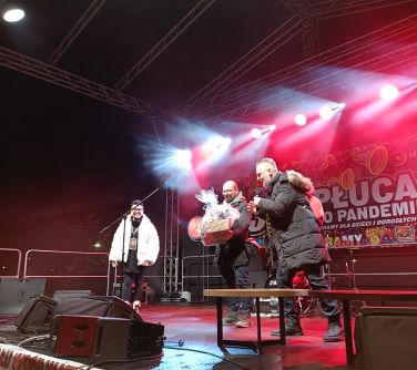 Na oświetlonej scenie trzy osoby. Mężczyzna przemawia do mikrofonu. Po prawej stronie stolik