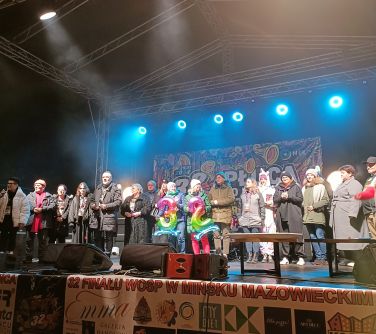 Na scenie grupa osób. Po środku trzymają balony kolorowe cyfry 32